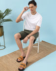 Man sitting on a chair wearing a white t-shirt, green shorts, and orange slides with a blue background