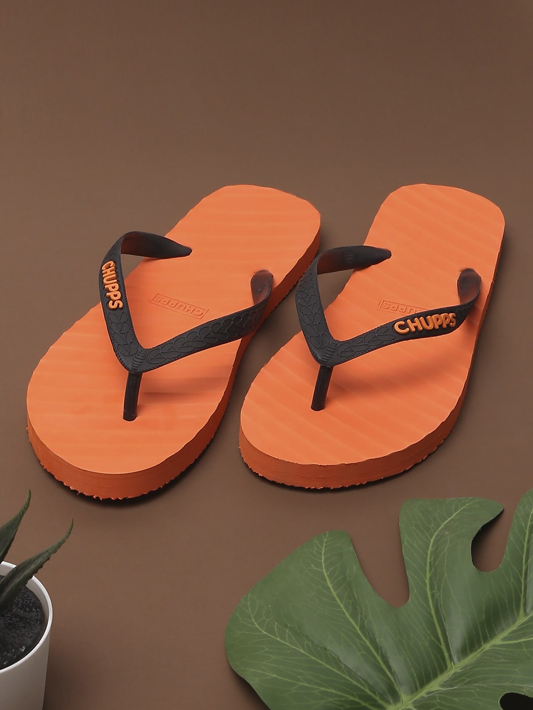 Chupps orange flip-flops with black straps for men, eco-friendly and stylish open-toe summer footwear on a tropical-themed background.