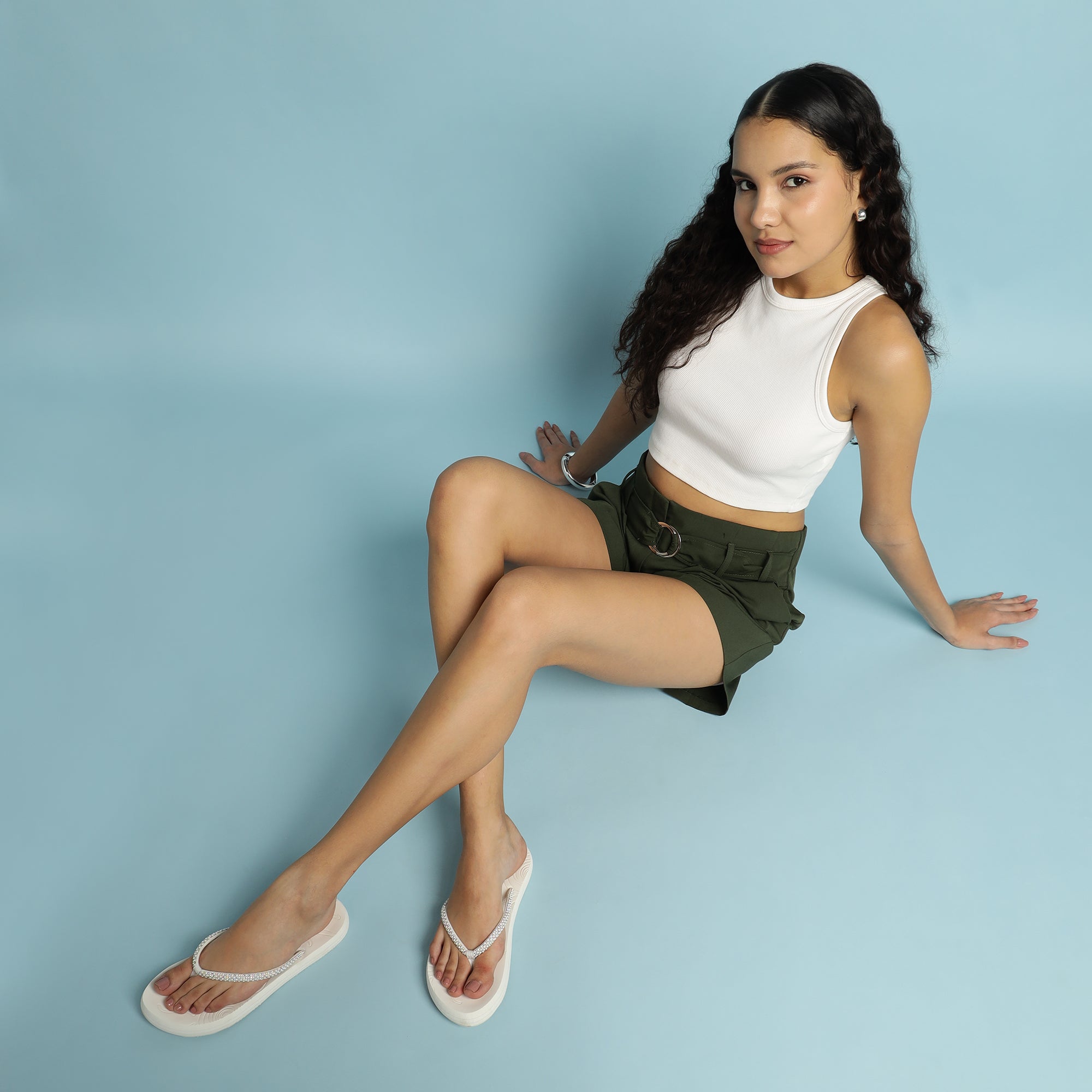 Woman sitting on a light blue background wearing a white sleeveless top and green shorts.