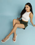 Woman sitting on a light blue background wearing a white sleeveless top and green shorts.