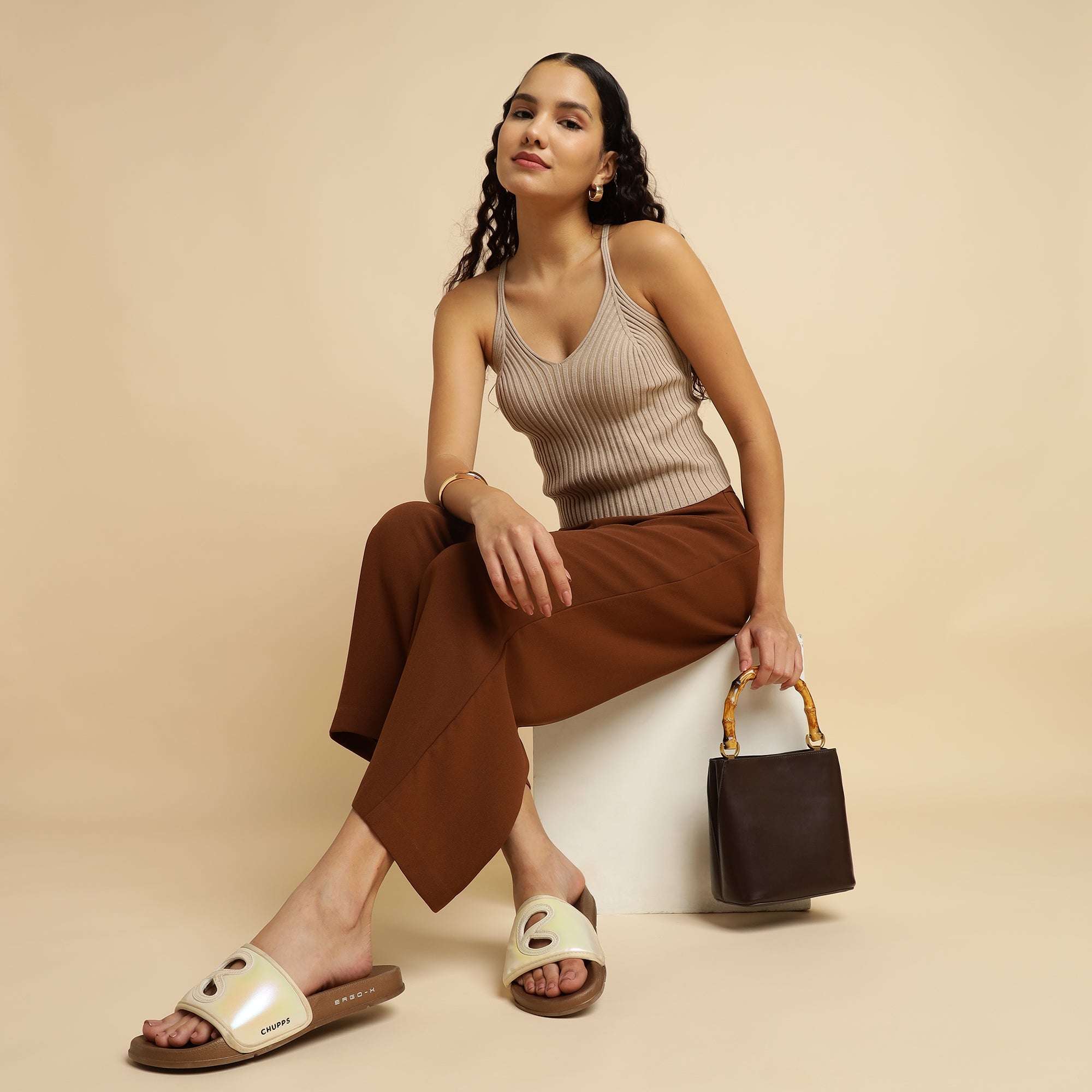 Woman sitting on a white stool holding a brown handbag against a beige background