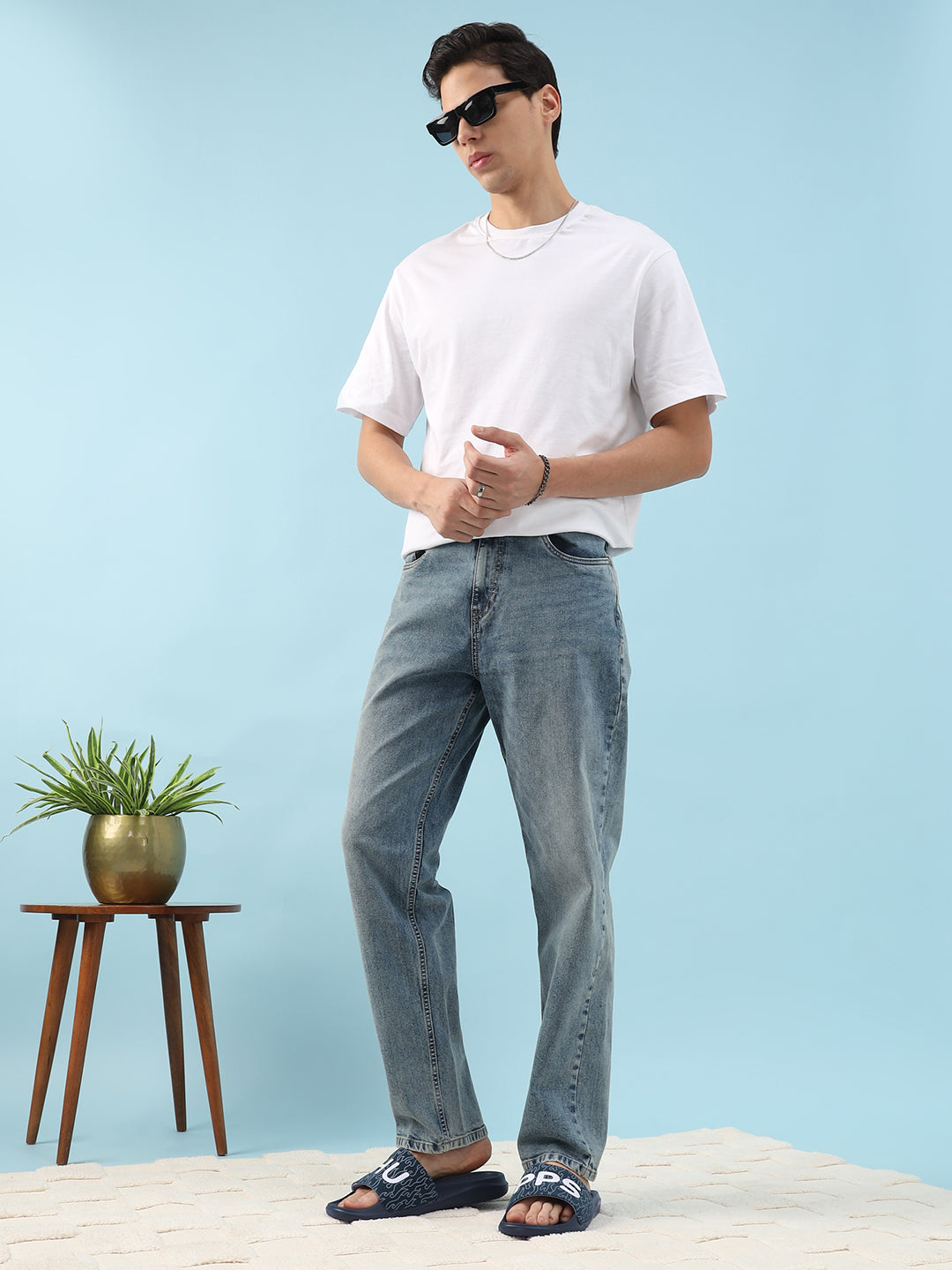 Man wearing a white t-shirt and blue jeans standing against a light blue background with a small plant and wooden stool.