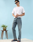 Man wearing a white t-shirt and blue jeans standing against a light blue background with a small plant and wooden stool.